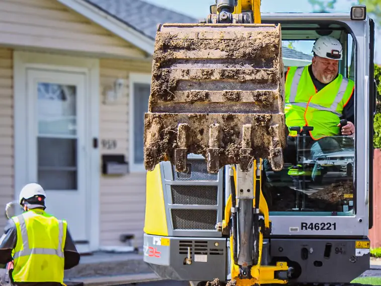 Legendary Service crew in hard hats running a compact excavator for a sewer line dig in a front yard